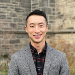 A person in a herringbone jacket and plaid shirt stands in front of a stone building, smiling with the confidence of an MPH graduate.